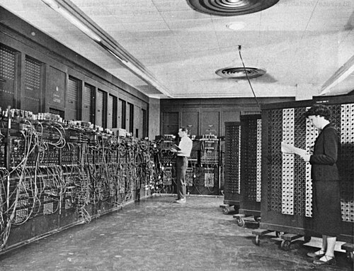Fotografia em preto e branco de um homem e uma mulher ajustando painéis cheios de cabos e chaves do computador ENIAC.