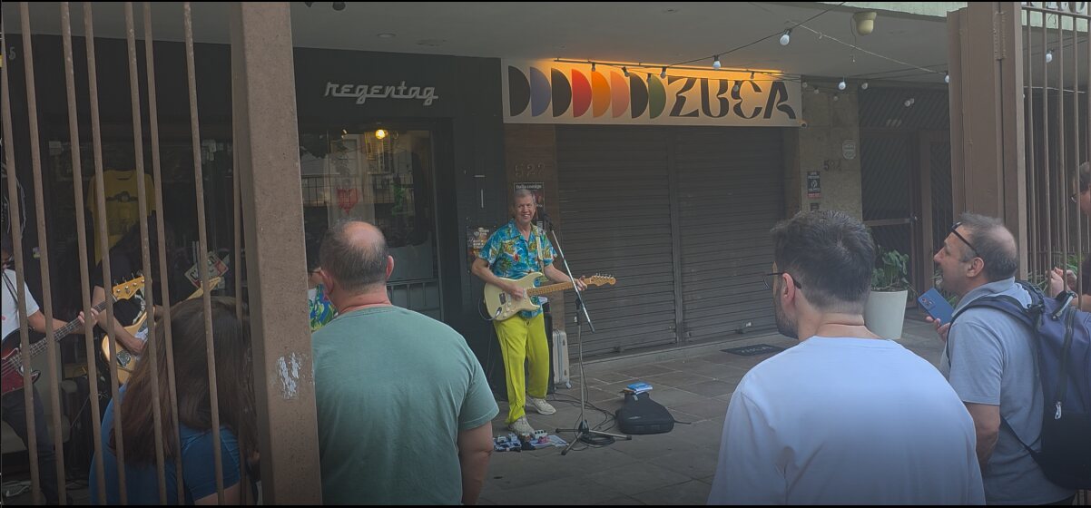 Wander Wildner, de camisa florida azul e calça amarela, toca guitarra na calçada da Rua João Telles, cercado por fãs que assistem e filmam com o celular